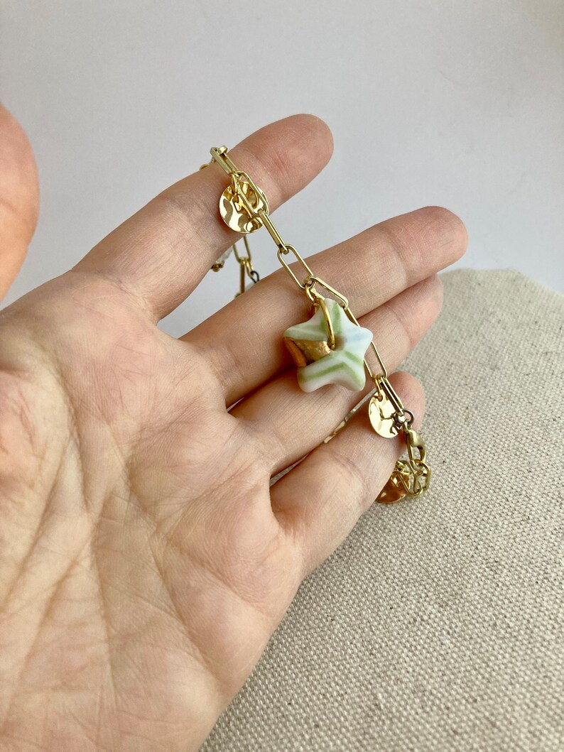 Pu&ograve; includere: Bracciale a catena dorata con un ciondolo a forma di stella a strisce verdi e bianche e dettagli dorati. Il bracciale include anche piccoli ciondoli rotondi dorati. Il gioiello &egrave; presentato su uno sfondo neutro.