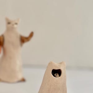 May include: A small, beige, cone-shaped ceramic sculpture with a cat-like silhouette and a circular opening revealing a tiny black and white figure inside. In the background, a taller, out-of-focus beige ceramic cat figure stands upright.