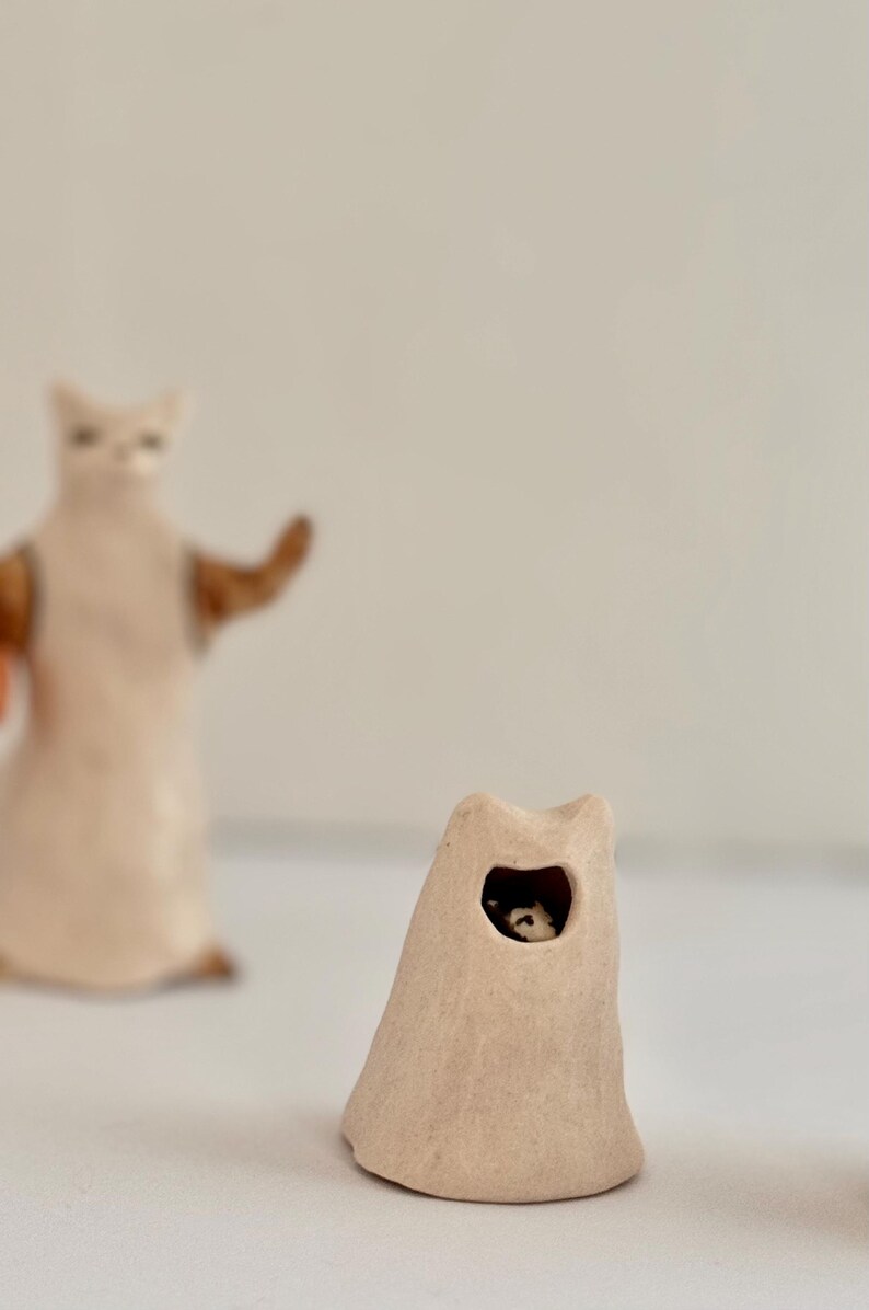 May include: A small, beige, cone-shaped ceramic sculpture with a cat-like silhouette and a circular opening revealing a tiny black and white figure inside. In the background, a taller, out-of-focus beige ceramic cat figure stands upright.