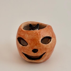 May include: A small, hand-sculpted ceramic pumpkin vessel with a glossy orange finish. The pumpkin features carved, black-painted eyes, a nose, and a wide, smiling mouth. A tiny, black-painted cat figure is visible peeking out from the top opening.