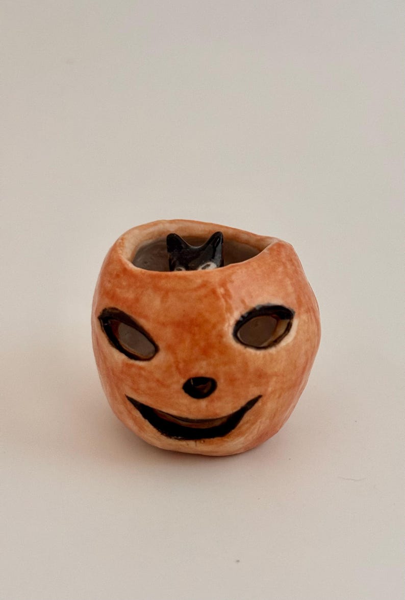 May include: A small, hand-sculpted ceramic pumpkin vessel with a glossy orange finish. The pumpkin features carved, black-painted eyes, a nose, and a wide, smiling mouth. A tiny, black-painted cat figure is visible peeking out from the top opening.