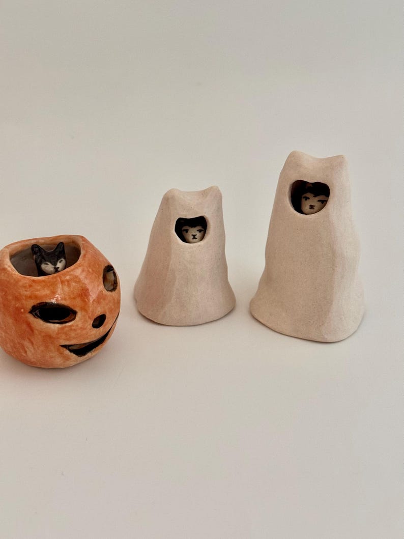May include: A ceramic orange pumpkin-shaped vessel containing a small black and white cat figure, alongside two off-white ghost-shaped ceramic figures, each featuring a small cat face peering through a cutout opening in the upper section.