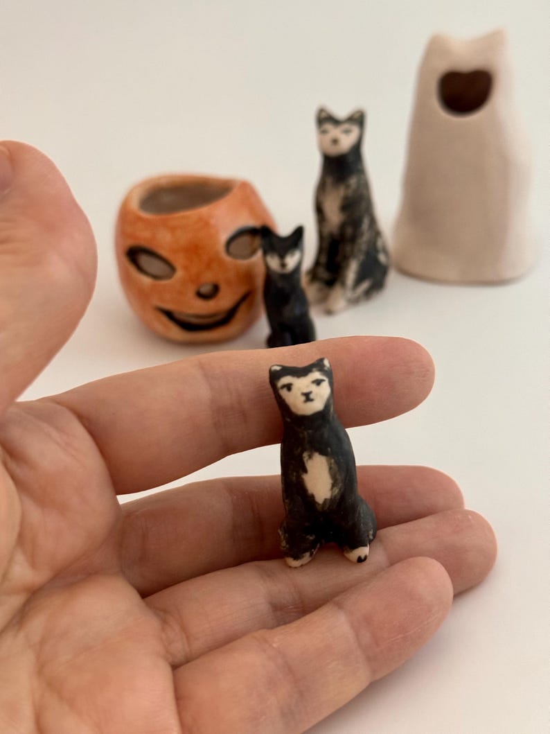 May include: A small black and white ceramic cat figurine with a painted face. In the background, there is an orange ceramic pumpkin-shaped container, a taller black and white ceramic cat figurine, and a pale beige ceramic ghost-shaped object.