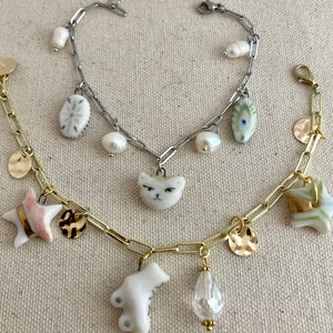 Pu&ograve; includere: Tre braccialetti con ciondoli vari. Un braccialetto &egrave; dorato con ciondoli a forma di stella, disco e orso. Un altro &egrave; argentato con ciondoli a forma di gatto, perla e ovale. Il terzo &egrave; dorato con ciondoli a forma di stella e disco. Lo sfondo &egrave; in tessuto neutro.