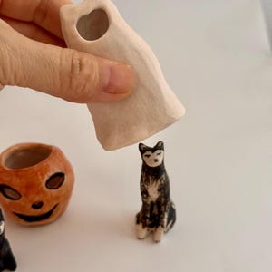 May include: A small, off-white ceramic ghost figurine with a hollow top, held above a miniature black and white cat figurine. To the left sits a small, orange, round ceramic pumpkin-shaped container with a carved face and dark eye sockets.