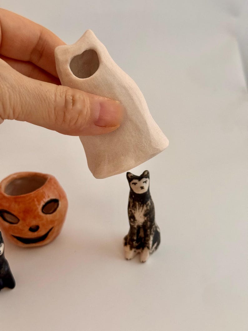 May include: A small, off-white ceramic ghost figurine with a hollow top, held above a miniature black and white cat figurine. To the left sits a small, orange, round ceramic pumpkin-shaped container with a carved face and dark eye sockets.