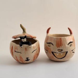 May include: Two ceramic containers with orange and cream glaze. The left container has a lid topped with a small black cat figure and a painted cat face. The right container features a wide, grinning cat face with two small pointed ears on the rim.