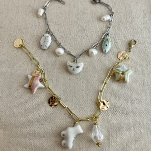 Pu&ograve; includere: Due braccialetti con ciondoli. Uno &egrave; argentato con un ciondolo a forma di gatto e perle, l'altro &egrave; dorato con ciondoli a forma di stella, moneta e goccia. I braccialetti sono su uno sfondo neutro.