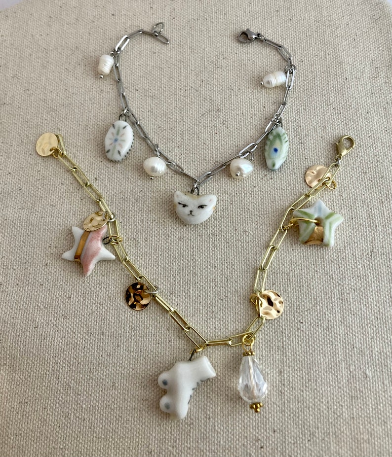 Pu&ograve; includere: Due braccialetti con ciondoli. Uno &egrave; argentato con un ciondolo a forma di gatto e perle, l'altro &egrave; dorato con ciondoli a forma di stella, moneta e goccia. I braccialetti sono su uno sfondo neutro.
