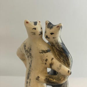 May include: A ceramic figurine featuring two cats standing upright and embracing each other. The cats are painted in off-white with grey and black brushstroke markings, featuring small red dots on their ears and simple black facial features.