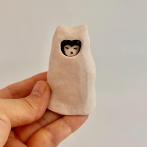 May include: A small, hand-sculpted ceramic figurine featuring a pale, matte-finish, cat-shaped hood or cloak. A tiny, detailed face with dark hair and painted features is visible through a circular opening in the center of the light-colored form.