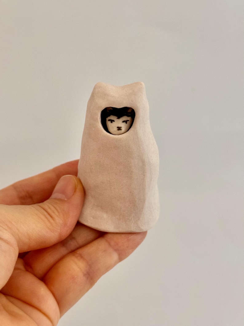 May include: A small, hand-sculpted ceramic figurine featuring a pale, matte-finish, cat-shaped hood or cloak. A tiny, detailed face with dark hair and painted features is visible through a circular opening in the center of the light-colored form.