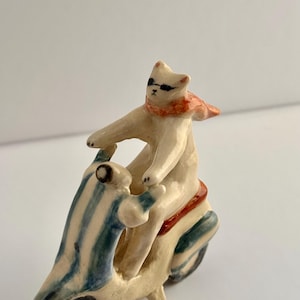 Peut inclure: Une petite figurine en céramique représentant un chat blanc portant des lunettes de soleil noires et une écharpe orange, conduisant un scooter crème avec des rayures bleues et blanches et une selle marron foncé.