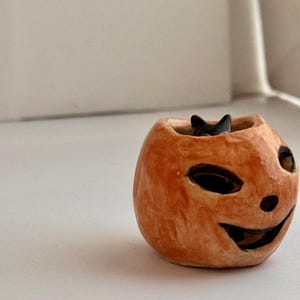 May include: A small, hand-sculpted ceramic pumpkin-shaped vessel with a glossy orange finish. The pumpkin features carved, dark-rimmed eye and mouth openings, and a small black cat figure is visible peeking out from the top opening of the piece.