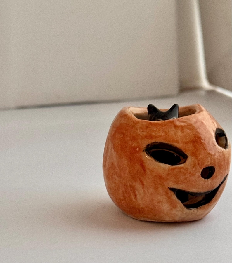 May include: A small, hand-sculpted ceramic pumpkin-shaped vessel with a glossy orange finish. The pumpkin features carved, dark-rimmed eye and mouth openings, and a small black cat figure is visible peeking out from the top opening of the piece.