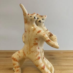 May include: A ceramic figurine depicting two orange tabby cats in a dancing pose. The cats have cream-colored bodies with orange patches and painted black facial features. One cat has an arm raised high while the other is held in a supportive embrace.