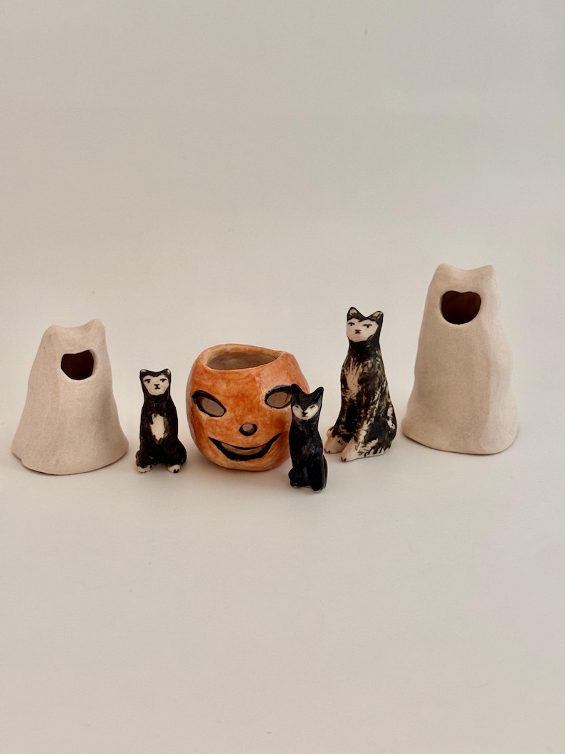 May include: A collection of five ceramic figurines including two off-white ghost shapes with cut-out eyes, two small black and white cats, and one central orange pumpkin-shaped vessel with a carved smiling face and dark eye openings.