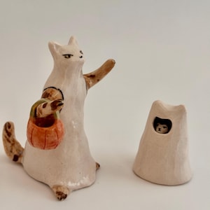May include: A ceramic figurine of a white cat wearing a ghost costume, holding an orange pumpkin bucket, with one arm raised. Beside it sits a separate white ceramic ghost hood with a small opening revealing a cat face peering out from inside.