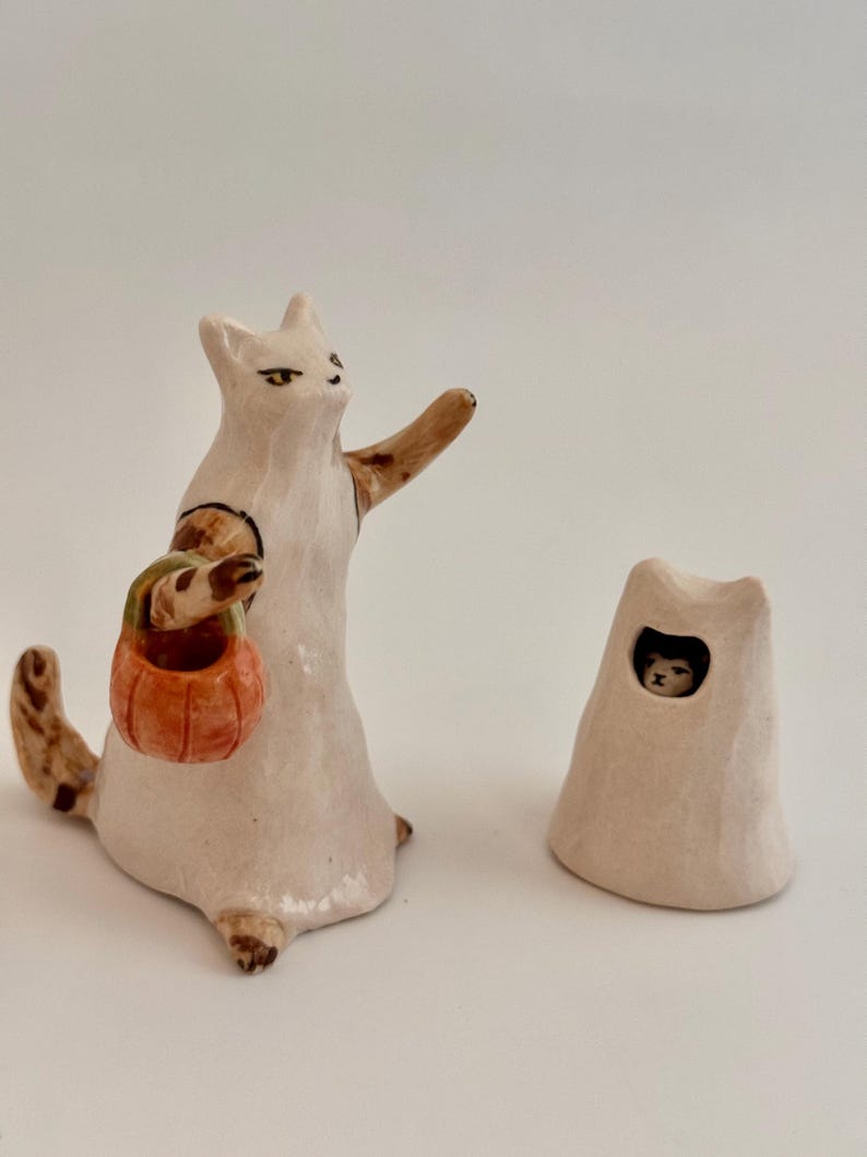 May include: A ceramic figurine of a white cat wearing a ghost costume, holding an orange pumpkin bucket, with one arm raised. Beside it sits a separate white ceramic ghost hood with a small opening revealing a cat face peering out from inside.