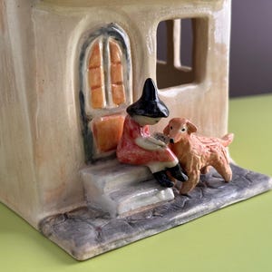 May include: A ceramic house figurine featuring a cream-colored structure with arched windows and a door. A figure wearing a black hat and red clothing sits on the front steps reading a book, accompanied by a small, textured golden-brown dog.