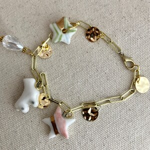 Pu&ograve; includere: Bracciale a catena dorata con ciondoli. I ciondoli includono un pattino a rotelle bianco, perline a forma di stella con dettagli verdi e rosa e ciondoli a disco dorati. Una perla trasparente a forma di goccia pende dal bracciale.
