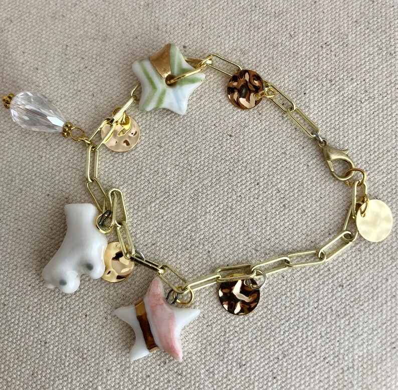 Pu&ograve; includere: Bracciale a catena dorata con ciondoli. I ciondoli includono un pattino a rotelle bianco, perline a forma di stella con dettagli verdi e rosa e ciondoli a disco dorati. Una perla trasparente a forma di goccia pende dal bracciale.