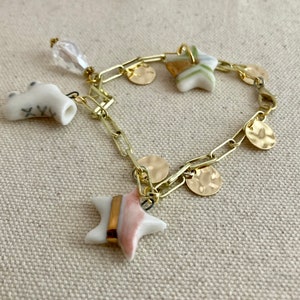 Pu&ograve; includere: Un braccialetto a catena color oro con pi&ugrave; ciondoli. I ciondoli includono piccoli dischi dorati, ciondoli a forma di stella con accenti rosa e oro e un ciondolo in ceramica bianca. Un ciondolo trasparente a forma di cristallo pende dalla catena.
