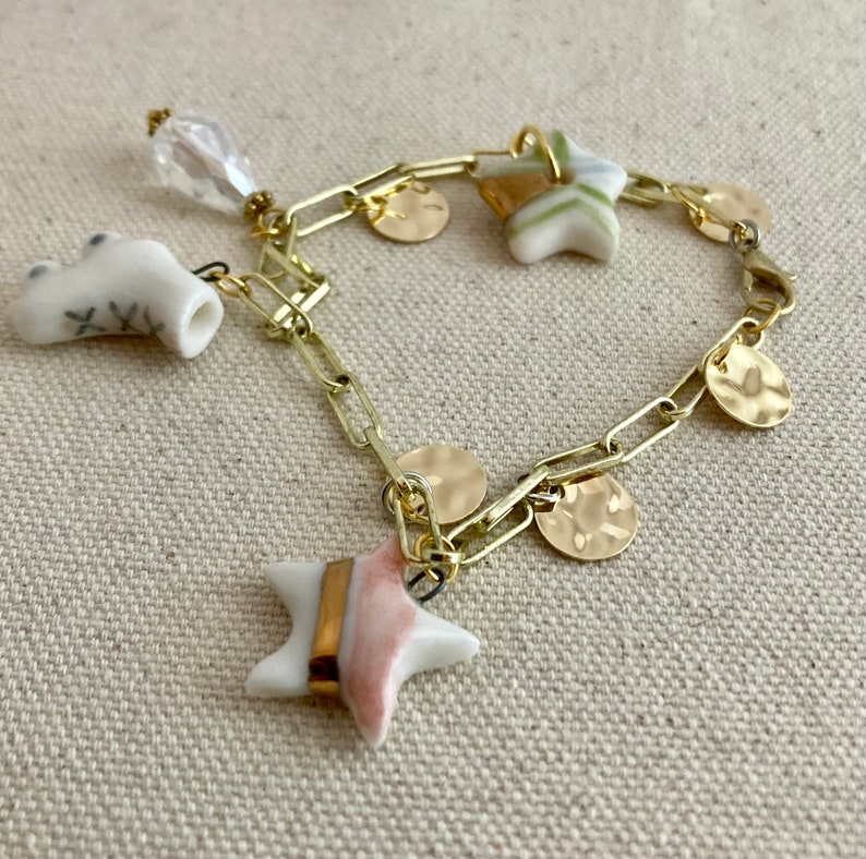 Pu&ograve; includere: Un braccialetto a catena color oro con pi&ugrave; ciondoli. I ciondoli includono piccoli dischi dorati, ciondoli a forma di stella con accenti rosa e oro e un ciondolo in ceramica bianca. Un ciondolo trasparente a forma di cristallo pende dalla catena.