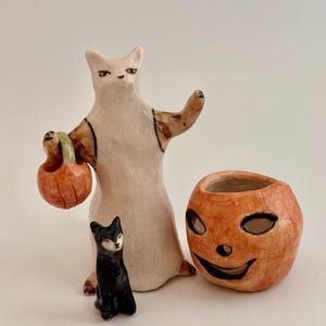 May include: A ceramic figurine of a white cat standing upright, holding a small orange pumpkin bucket, accompanied by a smaller black cat figurine and a separate orange ceramic pumpkin container with a carved jack-o'-lantern face.