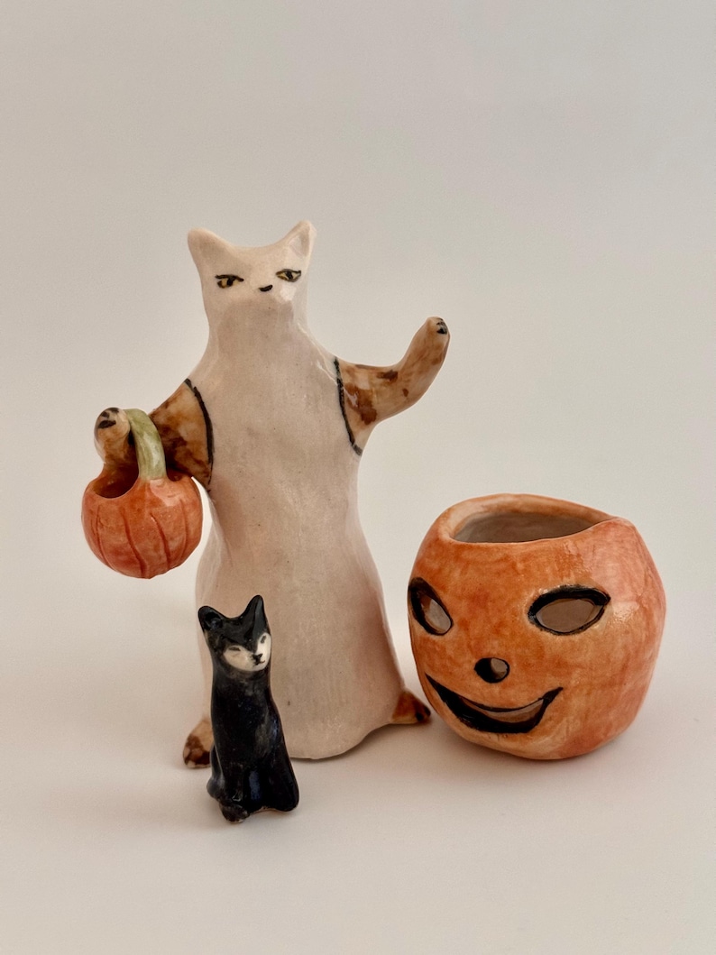 May include: A ceramic figurine of a white cat standing upright, holding a small orange pumpkin bucket, accompanied by a smaller black cat figurine and a separate orange ceramic pumpkin container with a carved jack-o'-lantern face.