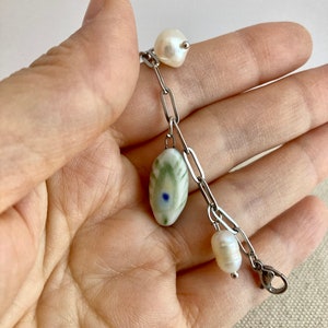 Pu&ograve; includere: Un braccialetto delicato con una catena d'argento, una perla ovale in ceramica e due perle d'acqua dolce. La perla in ceramica ha un motivo verde e bianco con un piccolo punto blu. Le perle sono bianche e di forma naturale.