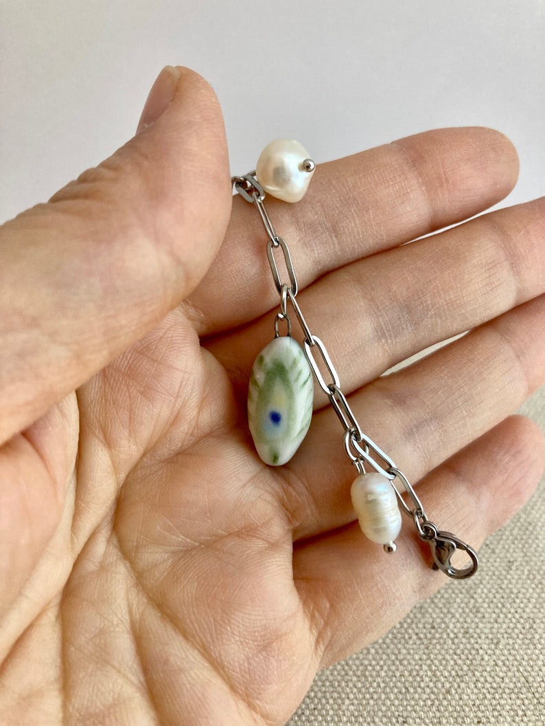 Pu&ograve; includere: Un braccialetto delicato con una catena d'argento, una perla ovale in ceramica e due perle d'acqua dolce. La perla in ceramica ha un motivo verde e bianco con un piccolo punto blu. Le perle sono bianche e di forma naturale.