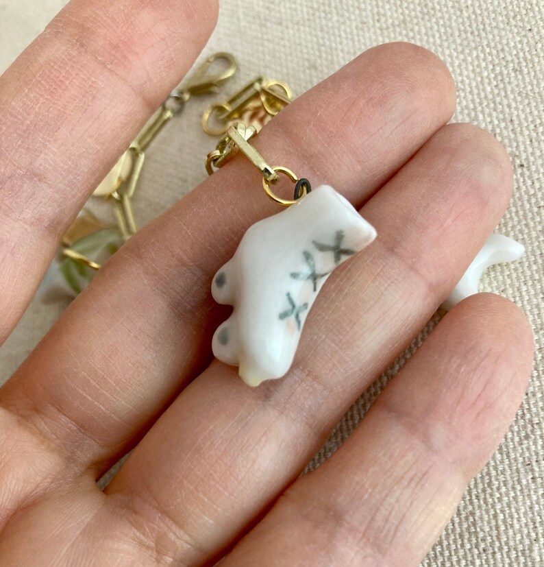 Pu&ograve; includere: Un piccolo ciondolo in ceramica bianca a forma di piede, con dettagli a punto croce neri. Il ciondolo &egrave; attaccato a una catena color oro con un moschettone. Il ciondolo &egrave; tenuto in una mano.
