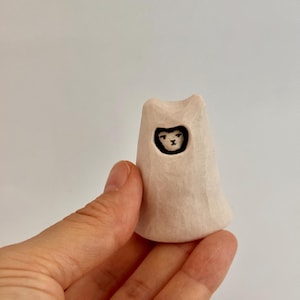 May include: A small, off-white, hand-carved wooden figurine shaped like a hooded figure. The figure has a smooth, matte texture and a carved opening at the top revealing a painted black and white face with simple, minimalist facial features.