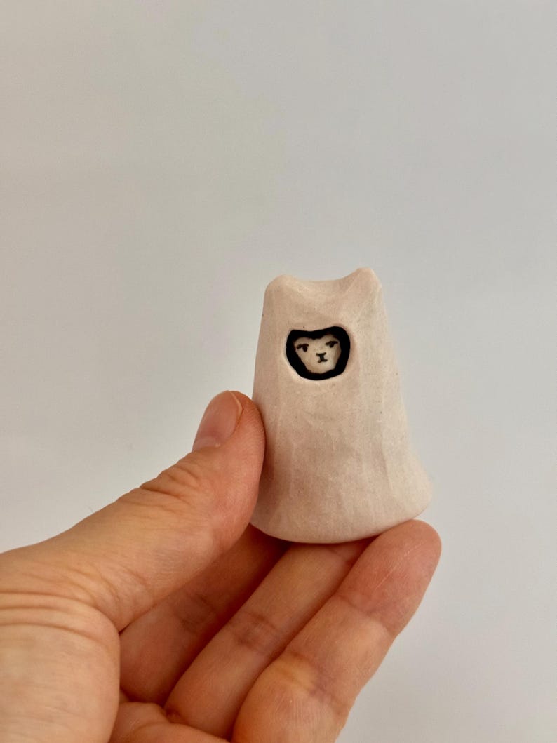 May include: A small, off-white, hand-carved wooden figurine shaped like a hooded figure. The figure has a smooth, matte texture and a carved opening at the top revealing a painted black and white face with simple, minimalist facial features.