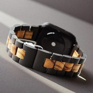 May include: A black smartwatch with a wooden band. The band alternates between dark black and light brown wood segments. The watch is angled, showing the back of the watch and the clasp.