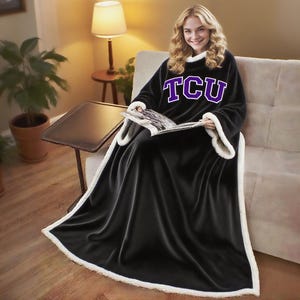 May include: A black wearable blanket with white trim and the letters "TCU" in purple. The blanket is made of a soft material and has sleeves. The person is sitting on a couch and reading a magazine.