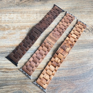 May include: Three wooden watch bands in varying shades of brown. The bands are made of rectangular wooden blocks. One band is dark brown, one is medium brown with the word "Engraved" on it, and the third is light brown.