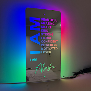 Könnte beinhalten: Eine Spiegel-Wanddekoration mit einem Regenbogen-Farbverlauf im Hintergrund und den Worten "I AM" in großen Buchstaben. Die Wörter "BEAUTIFUL, AMAZING, SMART, KIND, STRONG, FIERCE, CONFIDENT, POWERFUL, MOTIVATED, LOVED" sind vertikal unter den Worten "I AM" aufgelistet. Die Wörter "I AM" werden in kleineren Buchstaben unter der Wortliste wiederholt, gefolgt vom Namen "Alaska" in Kursivschrift.