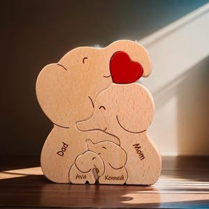 Peut inclure: Une figurine en bois d'une famille d'&eacute;l&eacute;phants avec un c&oelig;ur rouge. Les &eacute;l&eacute;phants sont empil&eacute;s les uns sur les autres, le plus grand en bas. Les &eacute;l&eacute;phants sont &eacute;tiquet&eacute;s "Papa", "Maman", "Ava" et "Kennedi".