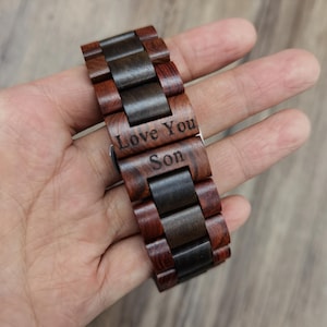 May include: Wooden watch with a dark brown and reddish-brown band. The watch band has the engraved text "Love You Son". The watch is made of wood and has a natural grain pattern.