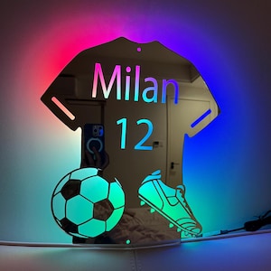 以下が含まれることがあります： サッカーをテーマにした装飾ミラー。ユニフォームの形をしており、「Milan」という名前と「12」の数字が表示されています。デザインにはサッカーボールとサッカーのクリートが含まれており、すべて鮮やかなマルチカラーLEDグローで照らされています。