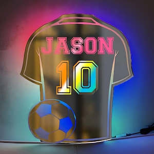 以下が含まれることがあります： 黒と金の鏡面加工されたサッカーユニフォームで、背中に「JASON」と「10」の番号が書かれています。手前に金のサッカーボールがあります。