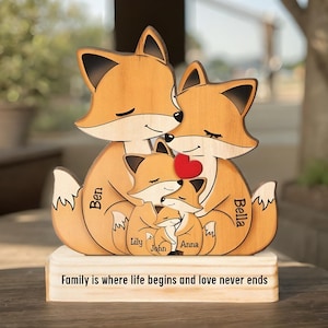 Puede incluir: Escultura de madera de una familia de zorros con un corazón. La escultura presenta dos zorros adultos y tres zorros más pequeños, con nombres como Ben, Bella, Lily, John y Anna. La base dice: "La familia es donde la vida comienza y el amor nunca termina."