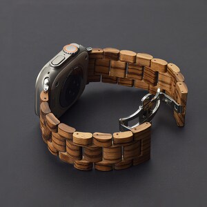 May include: A silver smartwatch with a wooden band. The band is made of rectangular wooden links with a striped pattern. The watch is open, showing the clasp. The background is a dark gray.