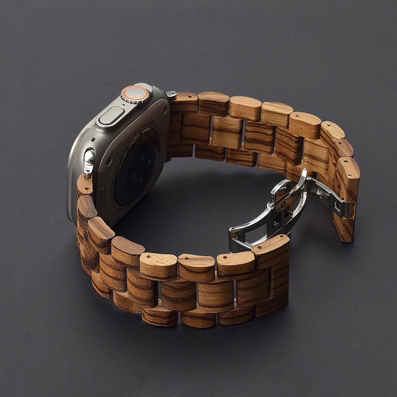 May include: A silver smartwatch with a wooden band. The band is made of rectangular wooden links with a striped pattern. The watch is open, showing the clasp. The background is a dark gray.