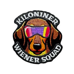 May include: An embroidered patch featuring a brown dachshund wearing purple and pink tinted goggles. The text KILONINER is arched above the dog, and the text WIENER SQUAD is arched below, both in white capital letters on a black background.