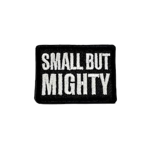 May include: A rectangular black embroidered patch with a stitched border. The text SMALL BUT MIGHTY is embroidered in white capital letters across the centre of the patch, featuring a clean, bold font style that stands out against the dark background.