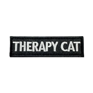 Peut inclure: Un écusson rectangulaire noir avec l'inscription "THERAPY CAT" brodée en blanc. Le patch a une bordure noire et est probablement fait de tissu.