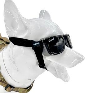 May include: Black protective eyewear for dogs featuring dark tinted lenses and a black adjustable strap. The frame displays the white text KILONINER on the side and lens. The glasses are positioned on a white, glossy, canine-shaped display head.